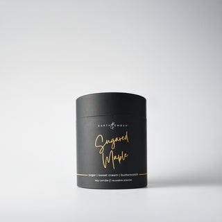 A black, cylindrical package containing a 'Sugared Maple' candle, with gold writing, indicating it is a soy wax candle from the Earth & Ember line