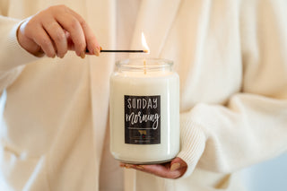 A hand lighting a 'Sunday Morning' candle with the person wearing a cream colored sweater as the background