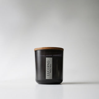 A black glass candle jar labeled 'Lucerno' with a wooden lid against a white background