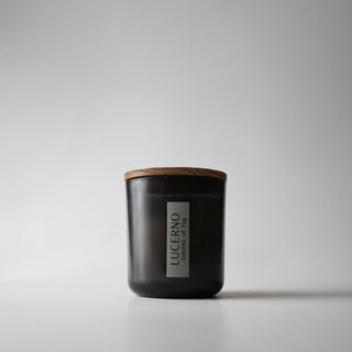 A black glass candle jar labeled 'Lucerno' with a wooden lid against a white background