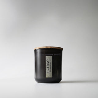 A black glass candle jar labeled 'Lucerno' with a wooden lid against a white background