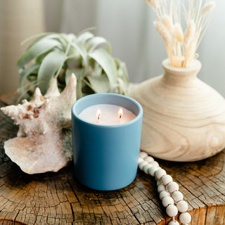 Lit candle in a blue container sitting on a wooden surface with a sea shell, vase and green plant in background