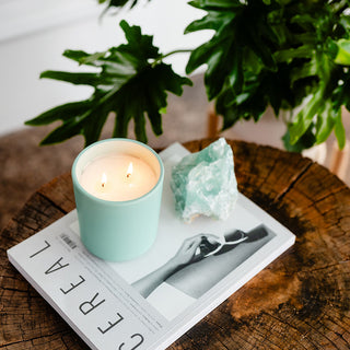 Lit candle in a seafoam green container on a wooded surface with a book and crystal 