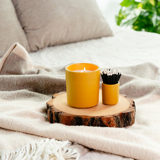Lit candle in a yellow container sitting on a wooden surface with a container of matches and bed in the background