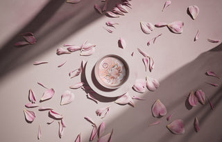 A light pink glass candle sitting on a saucer with pink flower petals scattered on a light pink background