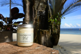 Candle labeled 'Saltwater Mist' on a wooden surface with a beach and palm trees in the background