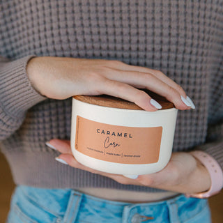 Person holding a 10 oz candle labeled 'Caramel Corn' with a wooden lid