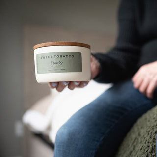 Person holding a 10 oz candle labeled 'Sweet Tobacco Leaves' in a casual setting